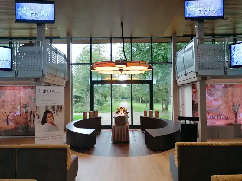 Rustig uitvaartcentrum met grote ramen en uitzicht op groen, comfortabele seating en informatief display over De Dageraad Uitvaartverzorging.